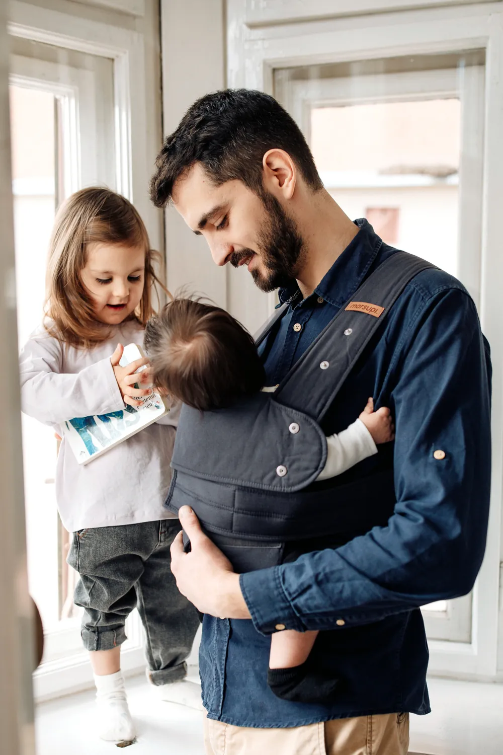 Vater trägt ein Baby in der Marsupi Classic 2 Babytrage in Grau, während ein Kleinkind das Baby füttert – Familienmoment am Fenster.