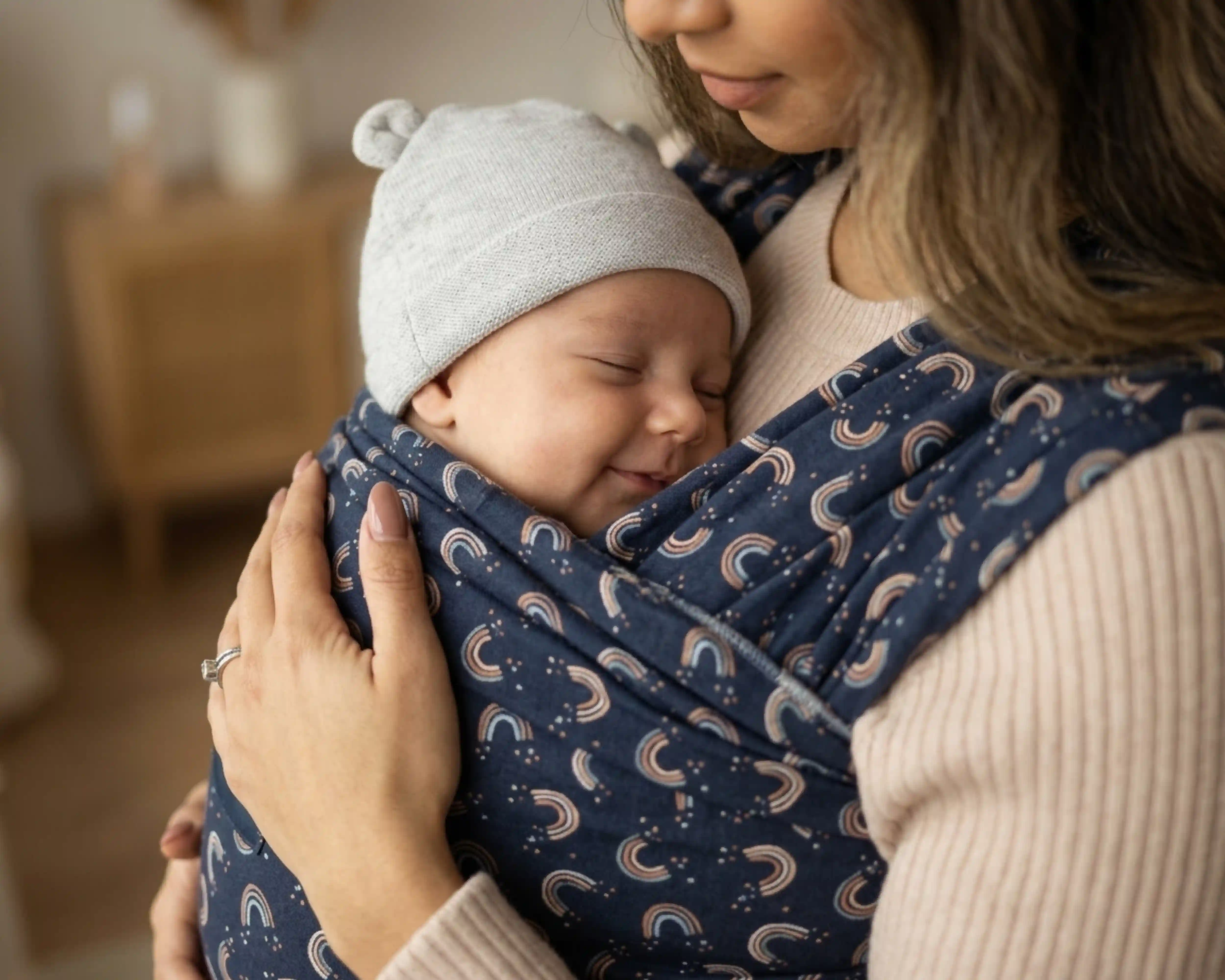 Nahaufnahme-Foto eines schlafenden Neugeborenen mit einer hellgrauen Bärenohrenmütze, das sicher in einem dunkelblauen gewebten Tragetuch mit einem Muster aus kleinen Regenbögen auf der Brust eines Elternteils schmiegt.