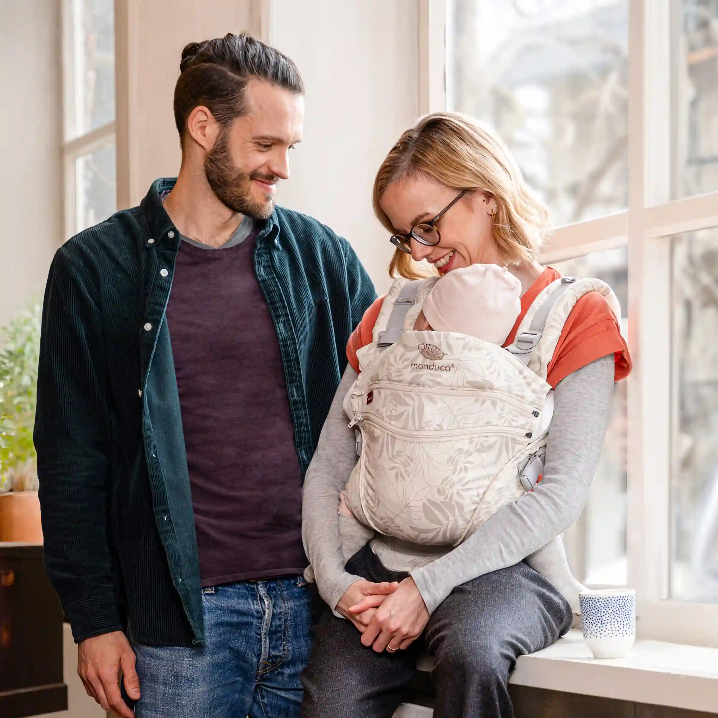 Mutter und Vater mit Baby in einer manduca® Trage, die sich lächelnd anschauen