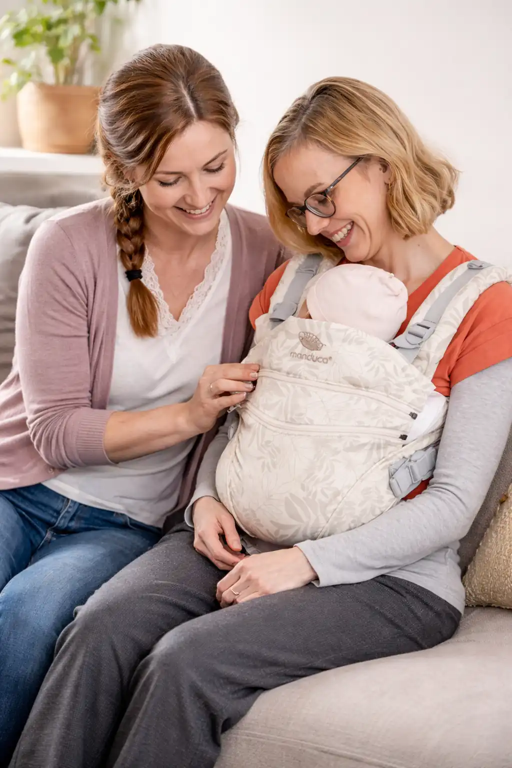 Hebamme sitzt neben einer Mutter auf dem Sofa und beide schauen gemeinsam auf eine angelegte ergonomische Babytrage mit Baby.