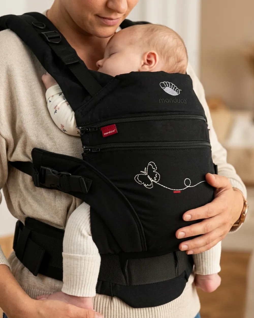 Frau in Strickpullover hält ein schlafendes Baby in einer schwarzen Manduca-Babytrage an ihrer Brust.