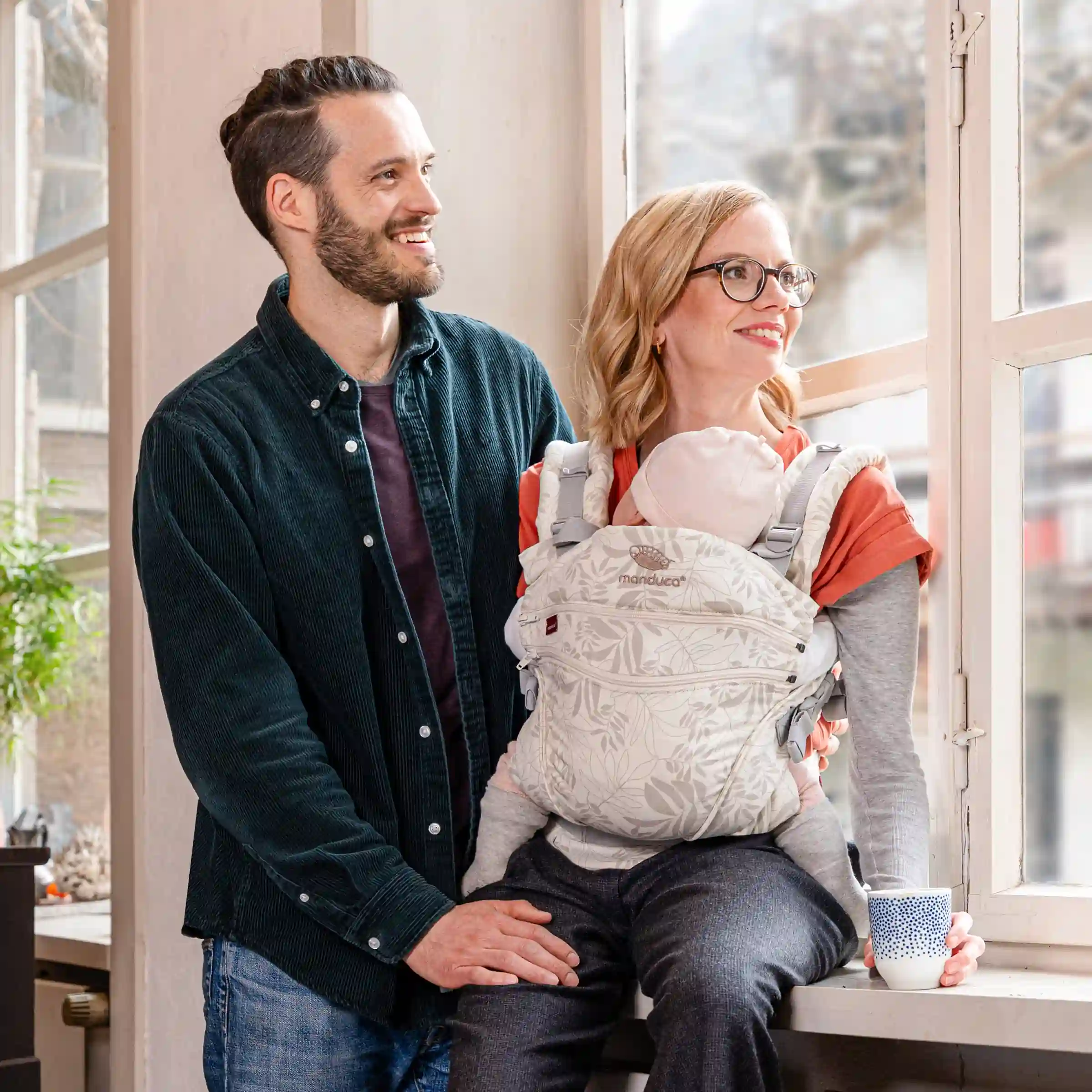 Eltern mit Neugeborenem in einer manduca® Babytrage sitzen entspannt am Fenster, das Baby kuschelt sich sichtbar eng an