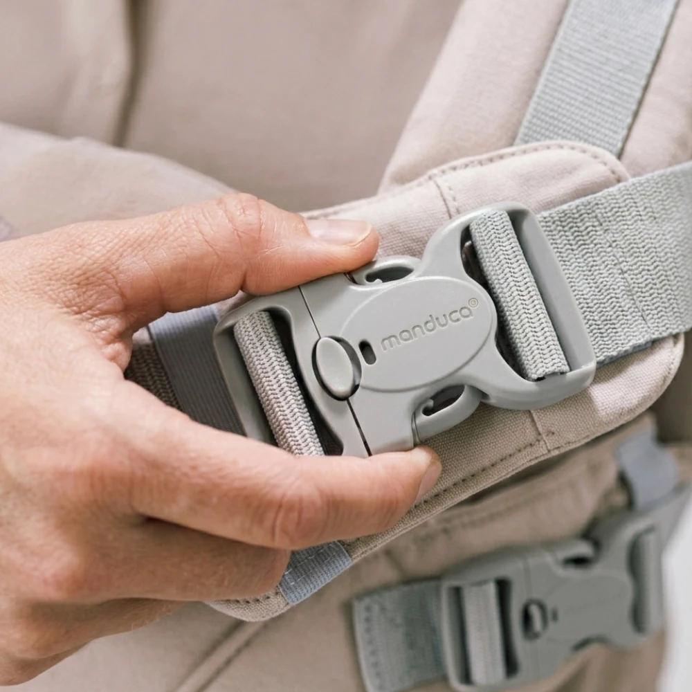 Nahaufnahme einer Hand, die eine graue Kunststoffschnalle mit dem eingeprägten Manduca-Logo an einer hellbeigen Babytrage schließt.